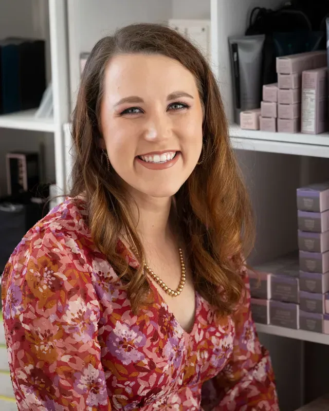 A smiling woman with long brown hair, wearing a floral blouse and gold necklace, posing in front of shelves of products.