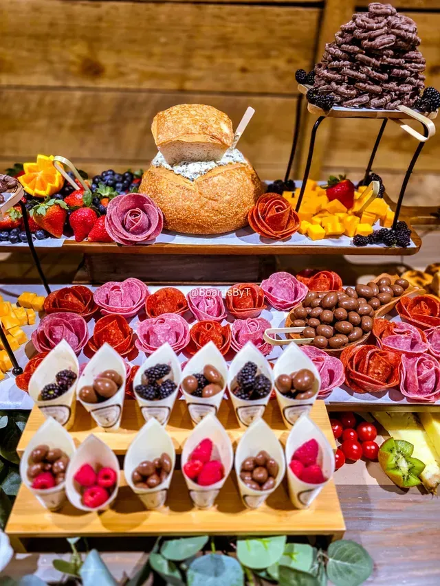 A rustic charcuterie display featuring a bread bowl, fruit, chocolate-covered treats, and individual snack cones.