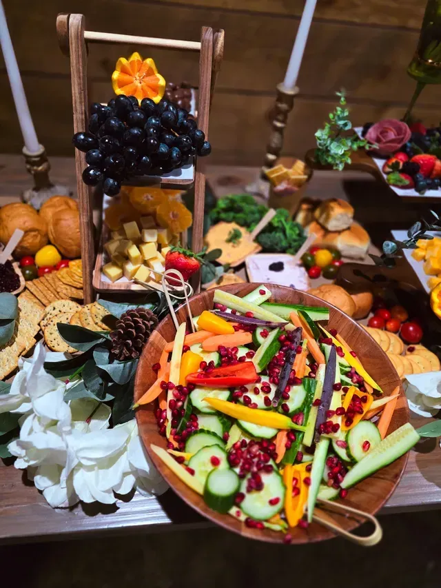 A wooden bowl of colorful salad with cucumbers, peppers, and pomegranate seeds sits on a table spread with food and decor.