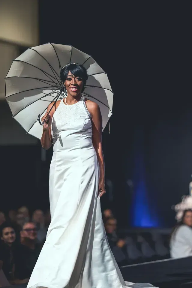 A person in a long white formal gown walks on a runway holding a white umbrella, smiling toward the audience.