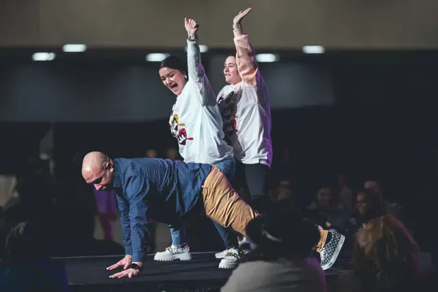 A man performs a push-up while two people stand behind him, cheering with arms raised on a stage before an audience.