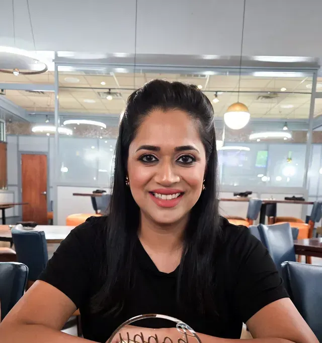 A smiling person with long dark hair, wearing a black shirt, sits at a table in a bright, modern restaurant setting.