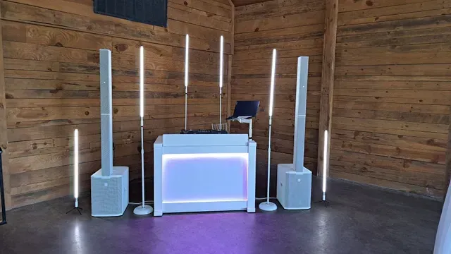 A white DJ booth with illuminated panels and tall speakers, set up on a concrete floor in a rustic wooden room.