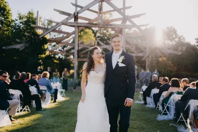 A newlywed couple smiling and walking together outdoors at a wedding ceremony with guests seated in the background.