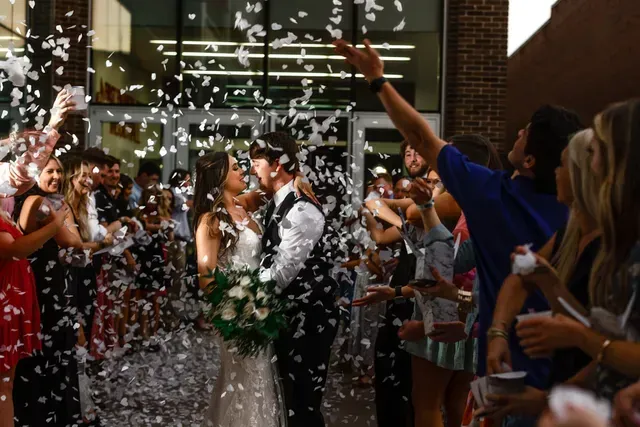 A bride and groom face each other as guests toss white confetti during a festive outdoor wedding exit.