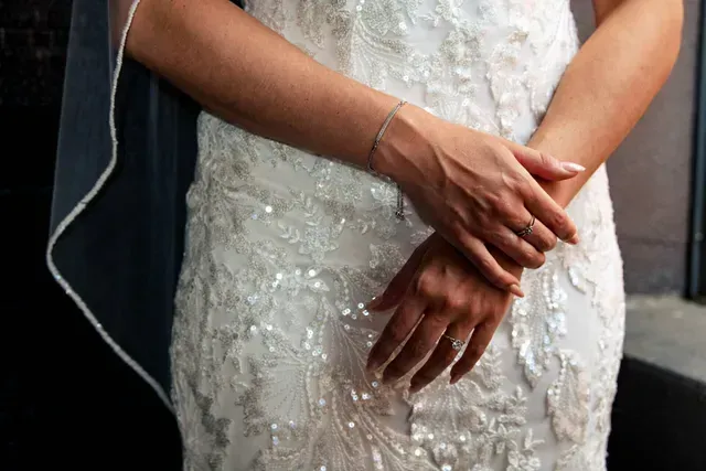 Hands clasped, wearing a sequined white lace wedding dress and rings.