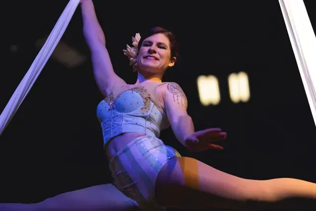 An aerialist in a light blue outfit performs a split while suspended by white fabric against a dark background.