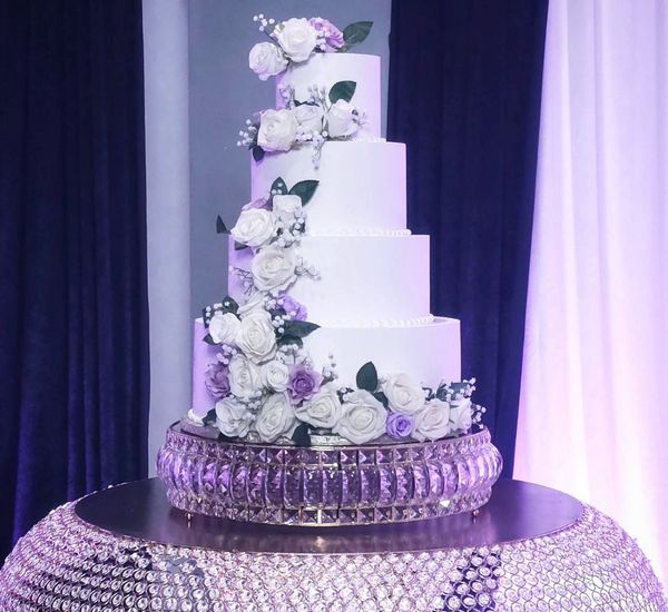 A four-tiered white wedding cake adorned with white and purple floral clusters, displayed on a crystal stand.