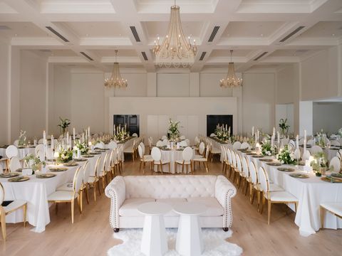 A spacious, elegant event hall with long tables, white linens, chandeliers, and a central white tufted loveseat.