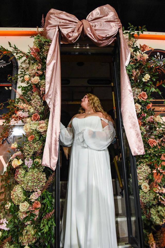 A person in a white wedding gown stands in a doorway decorated with lush flowers and a large, pink satin bow.