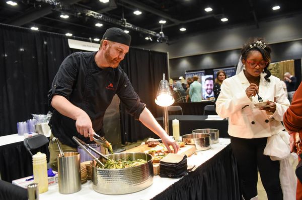 A chef in a black uniform prepares food from a metal bowl at a banquet, while a guest samples a portion nearby.