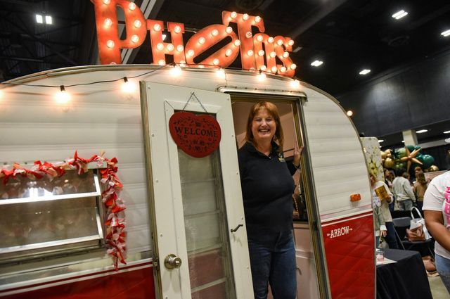 A smiling person stands in the doorway of a vintage, white-and-red camper trailer decorated with red, illuminated signage.
