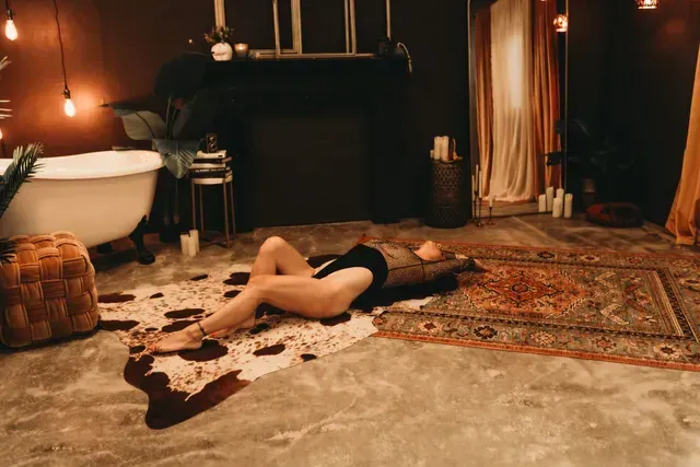 A person lies on a patterned cowhide rug in a dimly lit bathroom with a clawfoot tub, dark walls, and ornate rugs.