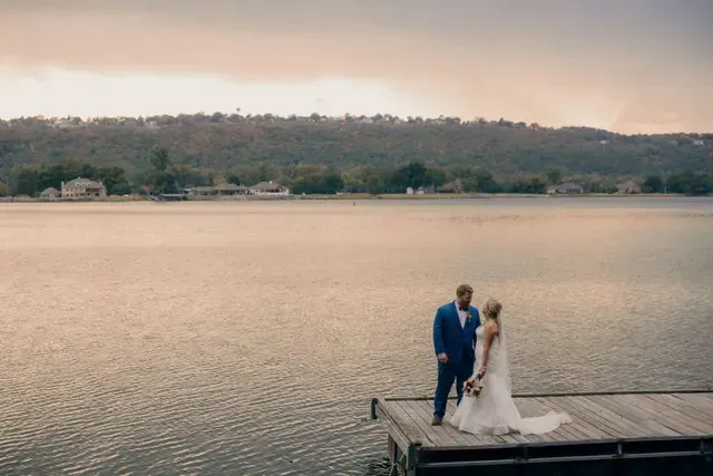 A bride and groom in wedding attire stand together on a wooden pier overlooking a calm, vast lake at sunset.