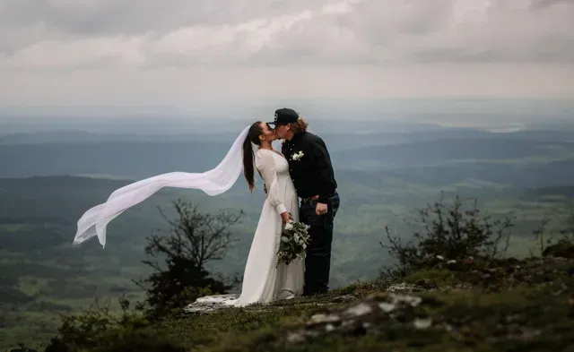 A couple in wedding attire kisses on a grassy clifftop, with the bride’s long veil blowing in the wind over a vast valley.