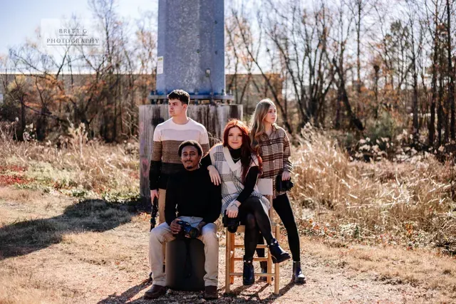 Four people pose for a photo in an outdoor field near a large pillar, with two sitting on stools and two standing.