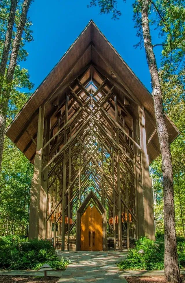 Tall, open-timber chapel nestled in a sunny forest, featuring intricate wooden beams and a central golden double door.