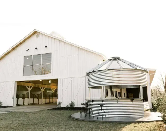 A white barn next to a round, corrugated metal silo converted into an outdoor bar with stools on a grassy lawn.
