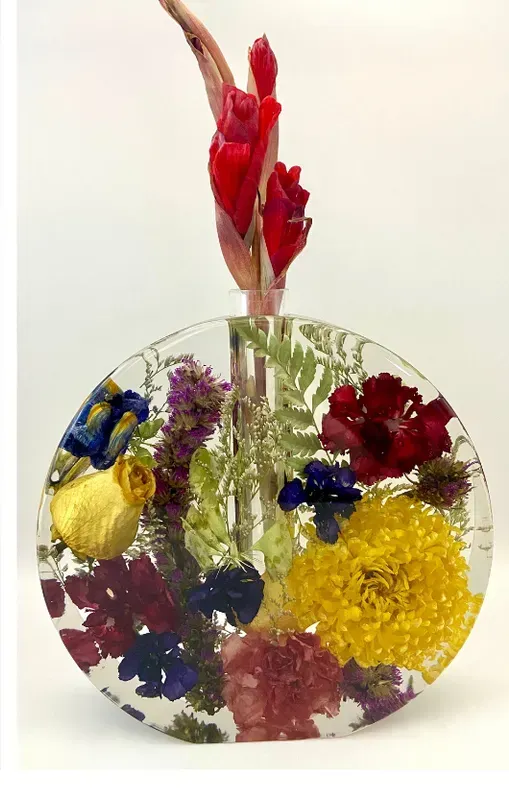 A circular resin vase filled with colorful pressed wildflowers, holding a single red ginger stalk in a central glass tube.