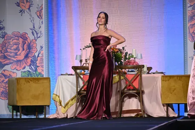 A person in a floor-length, dark red satin strapless gown poses on a stage in front of a dining table display.