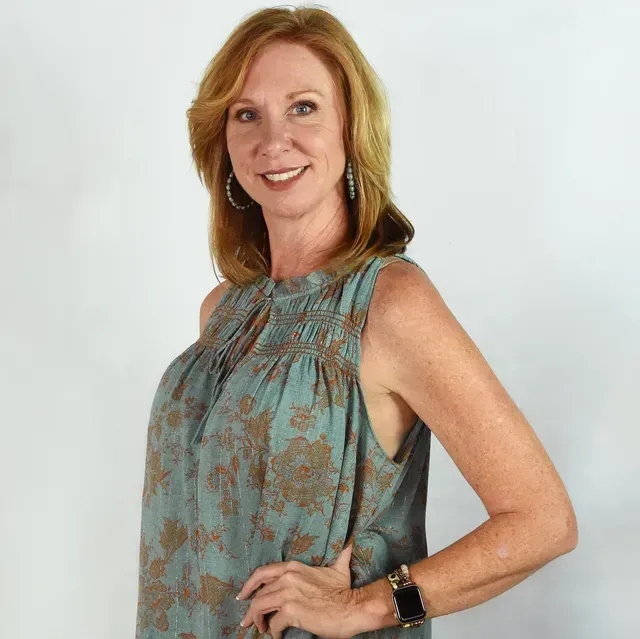 A person with shoulder-length auburn hair, wearing a green floral sleeveless top, poses against a plain white background.