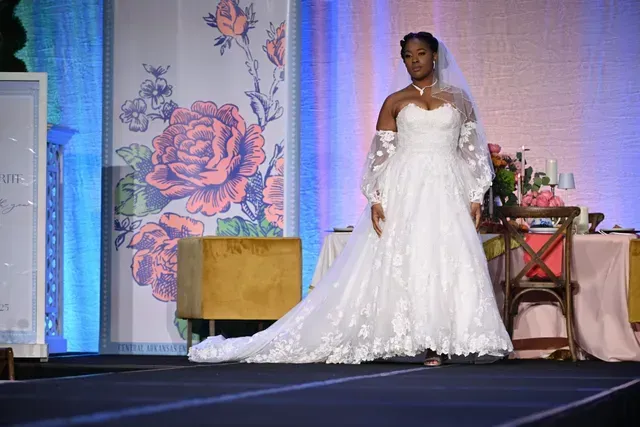 A model walks a runway wearing a white strapless wedding gown with lace details, sheer long sleeves, and a long train.