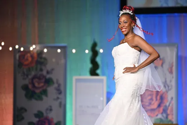 A smiling person in a strapless white mermaid wedding gown and veil poses on a stage decorated with floral panels.