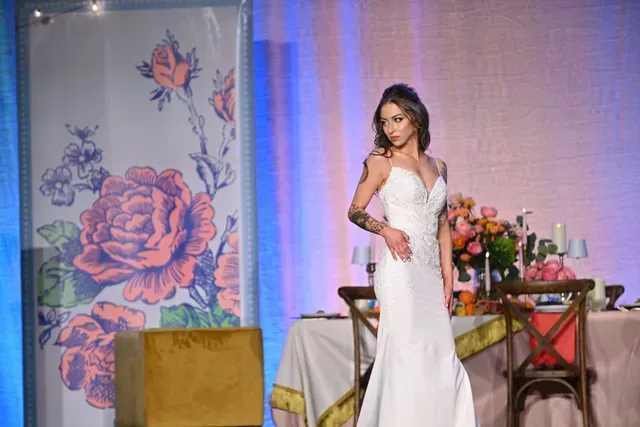 A person in a white gown posing in front of a rose-patterned screen with a draped table and wooden chairs.