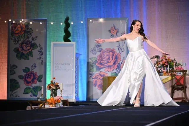 A person in a white wedding gown walks down a runway, arms outstretched, with floral panels in the background.