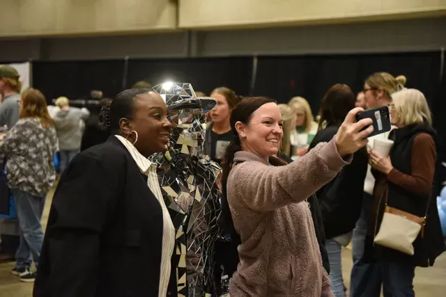 Two people pose for a selfie in an indoor exhibit space, one smiling and one looking toward a piece of wire-frame art.