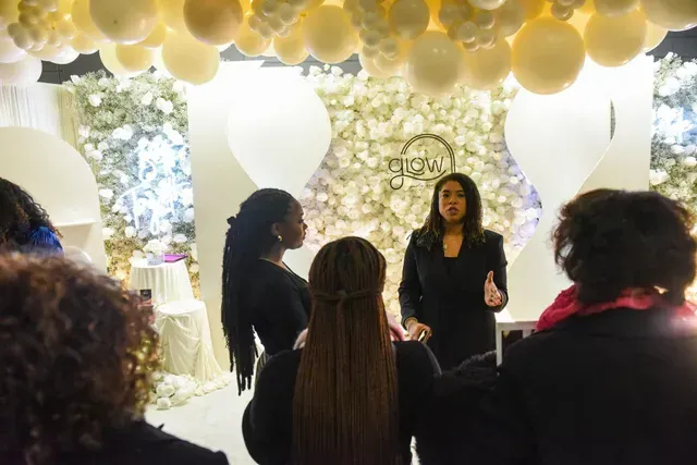 A woman in a dark suit speaks to a small group in a room decorated with white flowers and numerous beige balloons.