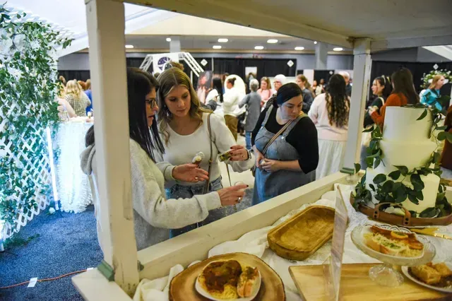 Two people look at food samples at an indoor wedding expo, with a tiered wedding cake visible on a table in the foreground.