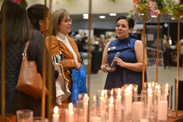 Three individuals converse at an indoor event, standing behind a table decorated with many lit candles in glass cylinders.
