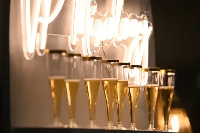 A row of champagne flutes filled with sparkling liquid, positioned in front of a glowing neon light sign.