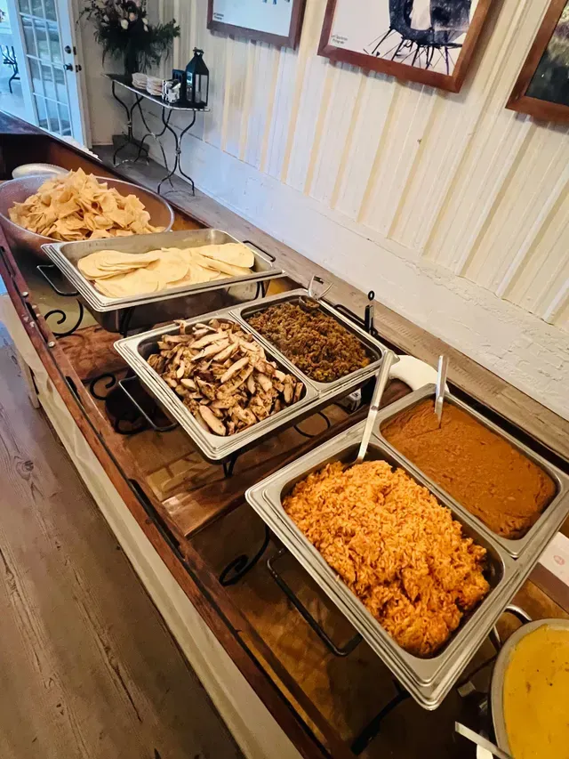 A buffet line featuring bowls of chips, tortillas, sliced chicken, seasoned meat, Spanish rice, and beans in metal pans.