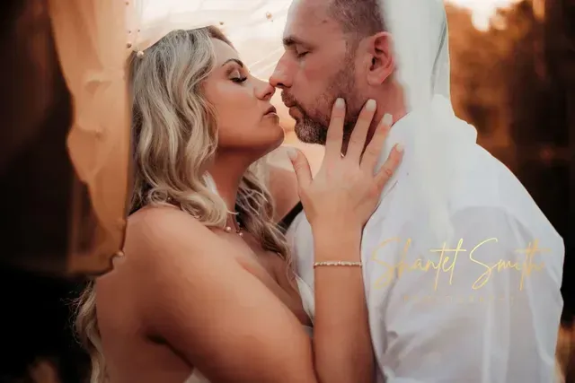 A couple in white attire embraces intimately under a draped fabric in an outdoor setting with warm lighting.