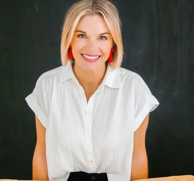 A smiling person with blonde bob hair wearing a white collared shirt and red earrings against a dark chalkboard background.