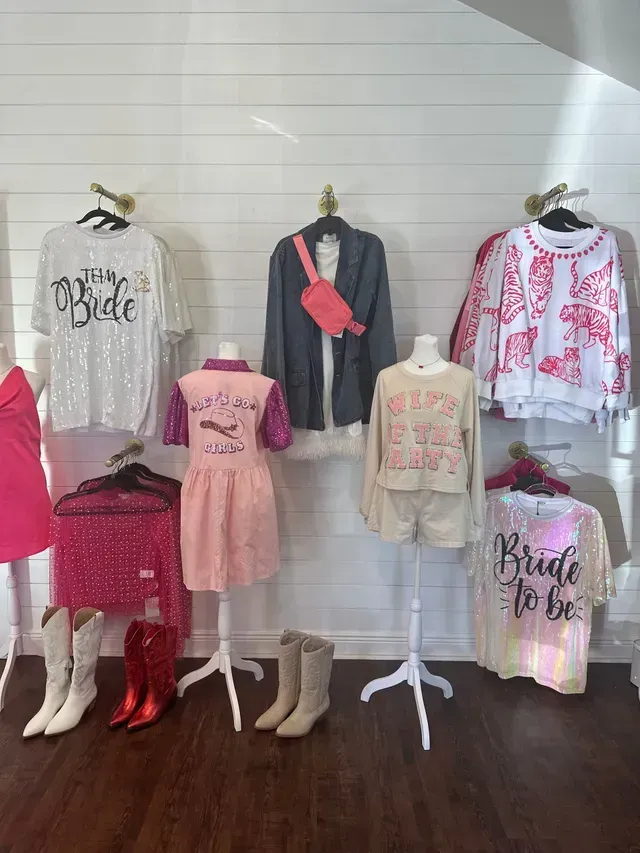 Clothing display in a boutique featuring white and pink bridal-themed shirts, sweaters, dresses, and cowboy boots.