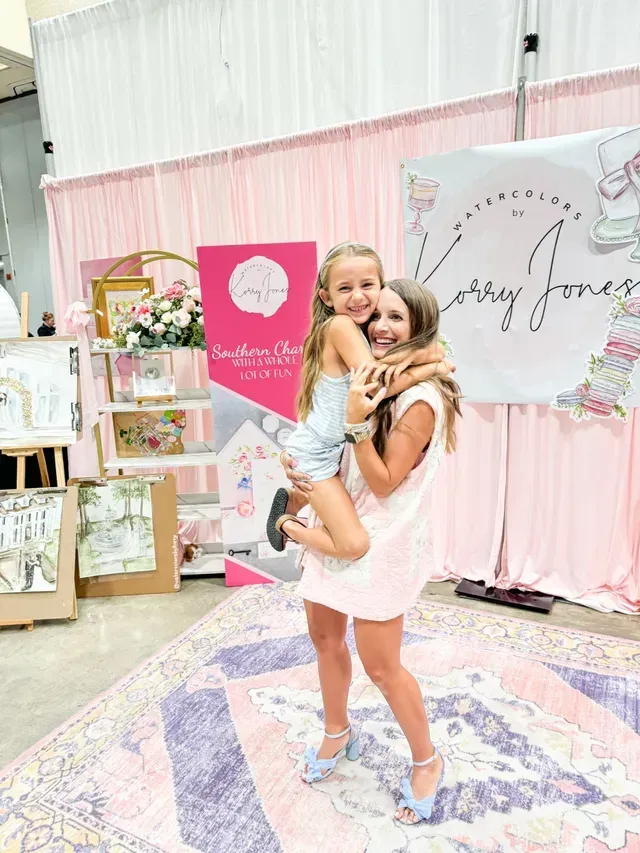 A smiling person holds a child in their arms in front of a pink-draped trade show booth with a branded sign.