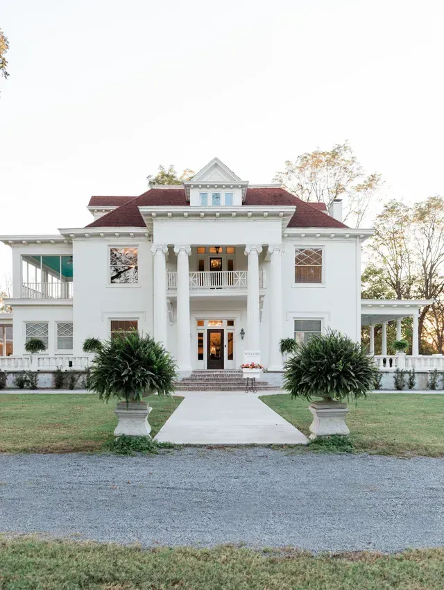 A white, two-story colonial-style mansion with a red roof, front columns, and a gravel driveway leading to the entrance.