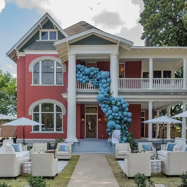 A red historic house decorated with a large blue balloon arch, featuring an outdoor seating area with chairs and umbrellas.