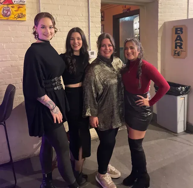 Four people smiling while standing in a line in a room with a bar sign.