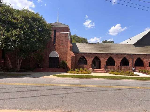 Red brick church building with a steeple, arched entrances, and a large tree in front under a blue sky.