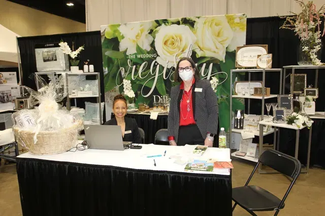 Two staff members stand at a booth promoting registry services, featuring a floral backdrop and various display items.