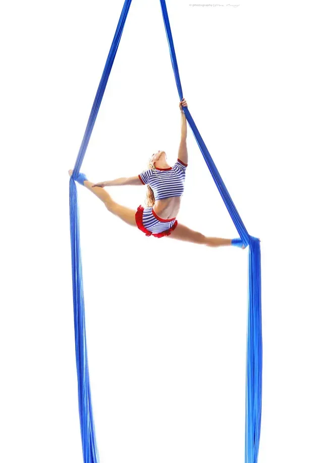 A performer in a blue-and-white striped outfit doing a split on blue aerial silks against a white background.