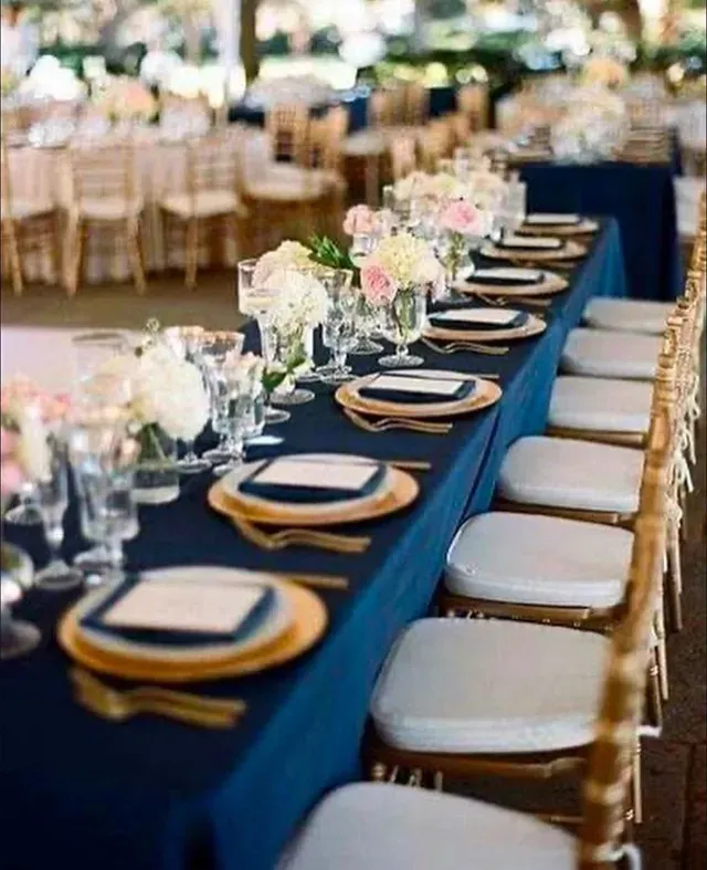 A long wedding table set with gold plates, navy linens, and pink and white floral centerpieces in a bright outdoor venue.