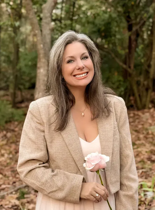 A smiling person in a tan blazer holds a single light pink rose in a wooded outdoor setting.