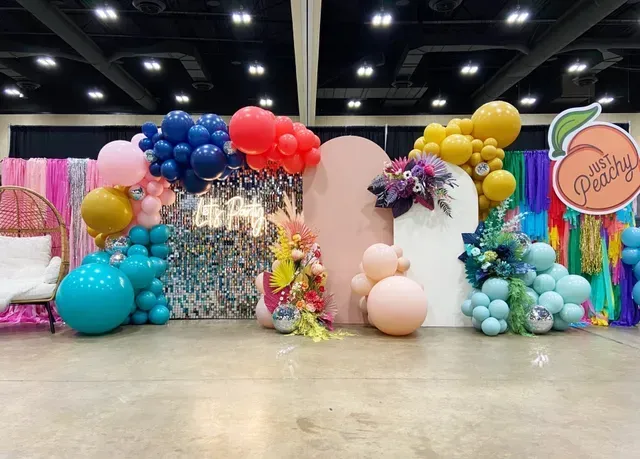 A vibrant balloon arch display featuring blue, red, gold, and peach tones with two arched panels and a 
