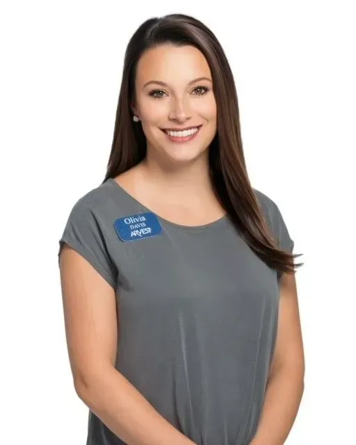 A smiling person with long dark hair wearing a gray top and a blue name tag against a white background.