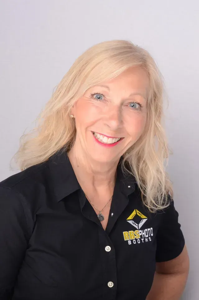 A smiling person with blonde hair wearing a black collared shirt with a yellow and white logo against a light background.